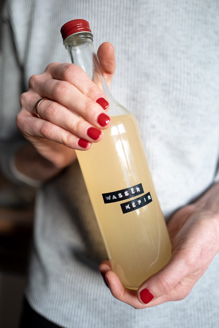 Hände mit rotlackierten Fingernägeln halten eine Flasche Wasserkefir